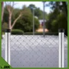 A close-up of a chain-link fence with two metal posts, set against a blurred outdoor background of trees and a building. Produktbild-Vorschau