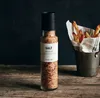 A bottle of chili blend salt next to a bucket of french fries on a wooden table. Produktbild-Vorschau
