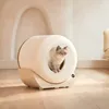 A cat sits inside a modern, self-cleaning litter box in a well-lit room with a light-colored wall and wooden floor. Produktbild-Vorschau