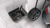 A black sink and a car tire are placed on a tiled floor in what appears to be a garage or workshop. Náhled obrázku produktu