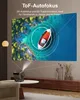 A projector is projecting an image of a boat on a body of water onto a wall. The projector is on a table in a living room setting. Produktbild-Vorschau
