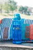 A blue Nalgene water bottle sits on a wooden surface in front of a striped bag, with a blurred outdoor background. Produktbild-Vorschau