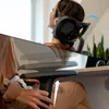 A woman is leaning back in an ergonomic office chair, with her head resting on the headrest. The image is split, showing the upper part of the chair and the woman's head and shoulders, and the lower part showing the armrest and a lever being adjusted. Produktbild-Vorschau