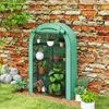 A green greenhouse filled with plants is placed against a brick wall in a garden setting. Voir les images du produit