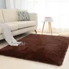 A brown shaggy rug is placed on a light wooden floor in front of a beige sofa. A striped blanket is draped over the sofa, and a decorative pillow with a tree pattern is on the sofa. A small white side table with books is to the right of the sofa. Náhled obrázku produktu