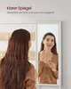 A woman is applying lipstick in front of a mirror, with a coat rack visible behind her. Náhľad obrázku produktu