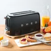 A black Smeg toaster is on a wooden table, surrounded by breakfast items like toast with jam, butter, and orange juice. The toaster has the 'SMEG' logo on it. Náhľad obrázku produktu