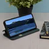 A dark green flip wallet case for a smartphone is displayed on a wooden desk, with a plant in the background and a notebook and pen to the side. Produktbild-Vorschau