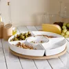 A white ceramic serving platter with multiple compartments filled with nuts and olives, resting on a wooden base. In the background, there are bottles of oil, salt and pepper shakers, a wedge of cheese, and grapes. Náhled obrázku produktu