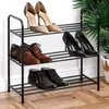 A black metal shoe rack with three shelves holding various pairs of shoes, placed indoors on a wooden floor. Náhled obrázku produktu