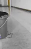 A bucket of black liquid is placed on a protective floor covering in a room with white walls. Produktbild-Vorschau