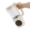 A hand holds a cream-colored automatic soy milk maker with a clear pitcher and a black control panel with gold trim. Náhled obrázku produktu