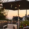 An outdoor patio scene at dusk, featuring a patio umbrella with string lights, a chair with a blanket, and decorative plants with lights. Produktbild-Vorschau