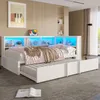 A bedroom scene with a white daybed pulled out with a trundle bed underneath, featuring a headboard with built-in shelves and blue LED lighting. A blanket and pillows are on the bed. A book is on the floor in the foreground. Náhled obrázku produktu