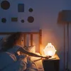 A woman in bed reaches for a night light on a bedside table. Produktbild-Vorschau