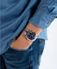 A person wearing a blue shirt and jeans, with a watch on their wrist. Produktbild-Vorschau