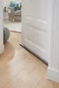 A door draft stopper is shown on a wooden floor in front of a white door, with a living room in the background. Podgląd zdjęcia produktu