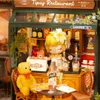 A miniature bistro scene with a doll behind the counter, a teddy bear at a table, and various food and drink items. Produktbild-Vorschau