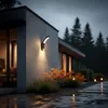A modern house exterior at dusk with an outdoor LED lamp illuminating the wall and surrounding plants. Anteprima dell’immagine prodotto