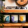 Produktfoto der "Climbing Mug Evo Keramiktasse mit Klettergriff" von YY Vertical; drei Tassen mit unterschiedlichen Griffen in einer Holzregal. Podgląd zdjęcia produktu