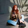 A young woman with red hair sits on the ground, holding a smartphone. She is wearing a trench coat and jeans, and is looking off to the side. The background is a modern architectural space with geometric shadows. Podgląd zdjęcia produktu
