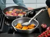 Two frying pans are on a stovetop, one with meat and vegetables, the other with fried food. A person's hands are visible, holding cooking utensils. Produktbild-Vorschau