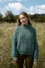 A woman stands in a field of tall grass and wildflowers, wearing a chunky knit sweater. Produktbild-Vorschau