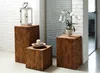 Three wooden pedestals of varying heights are arranged in a room with a white brick wall and a window with blinds. A lantern with a candle sits on the tallest pedestal, a bowl with a candle on the middle one, and a potted orchid on the shortest one. Voir les images du produit