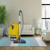 A yellow canister vacuum cleaner is positioned on a rug in a living room setting. Produktbild-Vorschau