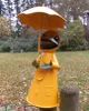 A garden decoration of a frog wearing a yellow raincoat and holding an umbrella, standing on a wooden post in a garden setting with fallen leaves. Produktbild-Vorschau