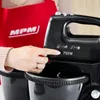 A person's hand is shown pressing a button on a black hand mixer. The mixer has speed settings indicated by icons. A red apron with the 'MPM' logo is visible in the background. Produktbild-Vorschau