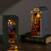 Two illuminated 3D wooden puzzle book nooks are displayed on a wooden surface, with one book nook resting on a large green book. Produktbild-Vorschau