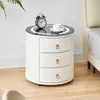 A white, round nightstand with three drawers and a glass top is shown next to a bed. An alarm clock and a cup of coffee are on top of the nightstand. Voir les images du produit