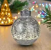 A decorative Christmas ornament with a winter forest scene and snowflakes, lit by internal LEDs, sits on a wooden surface. In the background, a blurred golden Christmas tree with lights and colorful string lights are visible. Produktbild-Vorschau