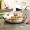 A stainless steel frying pan with food cooking inside, placed on a kitchen counter with ingredients like garlic and herbs nearby. Voir les images du produit