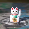 A white Maneki-neko cat figurine with red ears and a green collar sits on a graffiti-covered surface. Produktbild-Vorschau