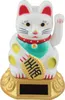 A white lucky cat figurine with yellow spots, a red collar, and a green scarf, holding a gold coin with Chinese characters. It is sitting on a gold base with a solar panel. Produktbild-Vorschau