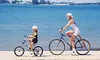 A woman and a child are riding bicycles along a waterfront path with a bridge and city skyline in the background. Podgląd zdjęcia produktu