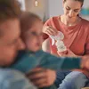 A woman is holding a breast pump and bottle while sitting with a baby and another person. Produktbild-Vorschau