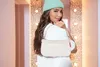 A young woman wearing a mint green beanie and a white t-shirt, holding a cream-colored fuzzy pouch bag over her shoulder. She is posing in front of a wall decorated with lights. Produktbild-Vorschau
