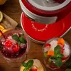 A close-up shot of a red and white ice crusher machine with three glasses of slushy drinks and fruit garnishes. Náhľad obrázku produktu