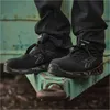 A person wearing black jeans and black safety shoes is sitting on a green metal surface. Náhľad obrázku produktu