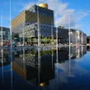 A modern building with a golden dome is reflected in a shallow pool of water, with tall white poles in the foreground. Voir les images du produit