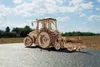 A wooden model tractor with a plow attached, placed on a dark surface with a blurred background of trees and a field under a cloudy sky. Náhľad obrázku produktu