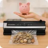 A pink piggy bank sits on a hand above a vacuum sealer with coins inside a vacuum-sealed bag. Podgląd zdjęcia produktu