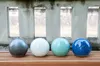 Four ceramic garden balls in different colors are arranged in a row on a wooden bench in front of a concrete wall with rectangular openings. Anteprima dell’immagine prodotto