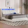 A modern white upholstered bed with a built-in LED headboard and USB ports, set in a bedroom with a rug and a potted plant. Produktbild-Vorschau