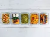 Five meal prep containers filled with different meals, arranged in a row on a white wooden surface. Anteprima dell’immagine prodotto