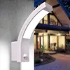 A white LED outdoor wall light with a motion sensor is mounted on a gray concrete wall, with a blurred background of a house at dusk. Produktbild-Vorschau