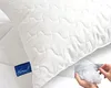 A close-up shot of a white pillow with a quilted pattern, showing a blue tag with a logo and text. An inset shows hands holding loose pillow filling. Produktbild-Vorschau
